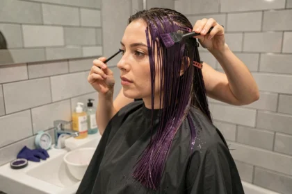 Femme appliquant une coloration violine foncée sur ses cheveux dans une salle de bain moderne avec des outils de coloration professionnels